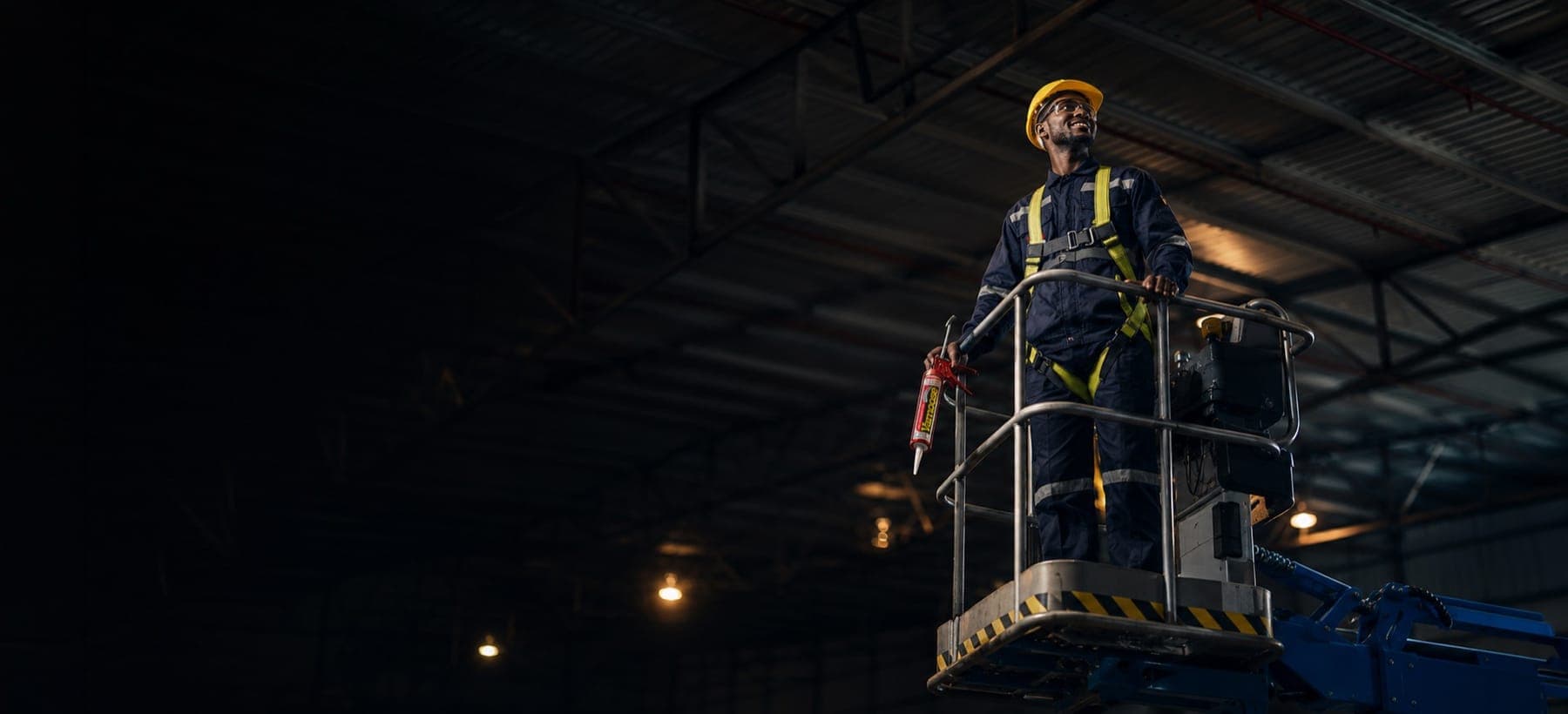 A BCS bird control technician on a cherry picker holding a Vamoose bird repellent gel cartridge at height inside an industrial warehouse